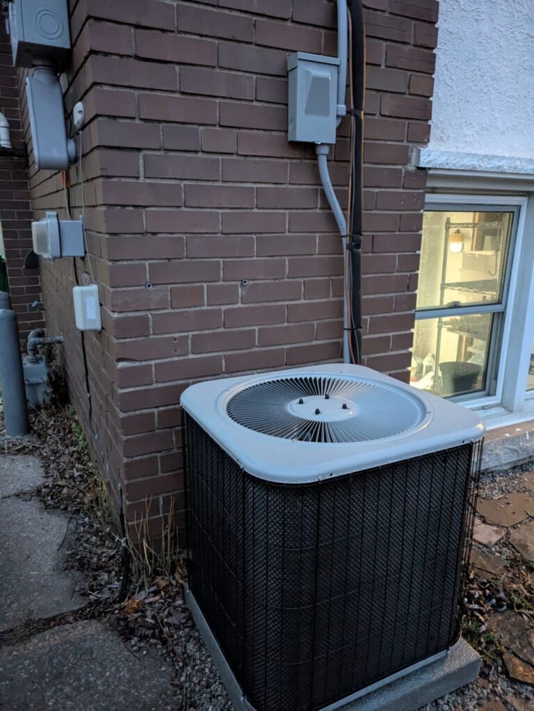 Air conditioner condenser with a black coil guard next to a brown brick wall, basement window and electrical disconnect