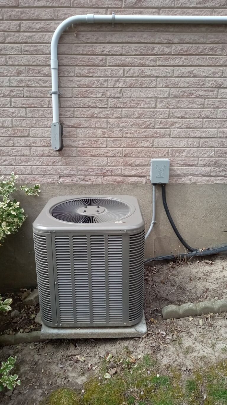 Grey air conditioner condenser with a top fan on a concrete pad against a light brick wall with conduit and a disconnect box