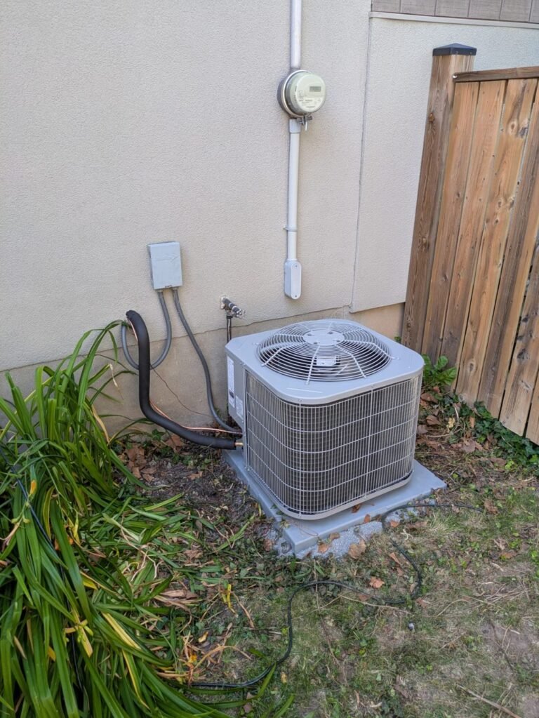 Air conditioner condenser installed in a narrow side yard between a stucco wall and a wooden fence with a disconnect and electric meter