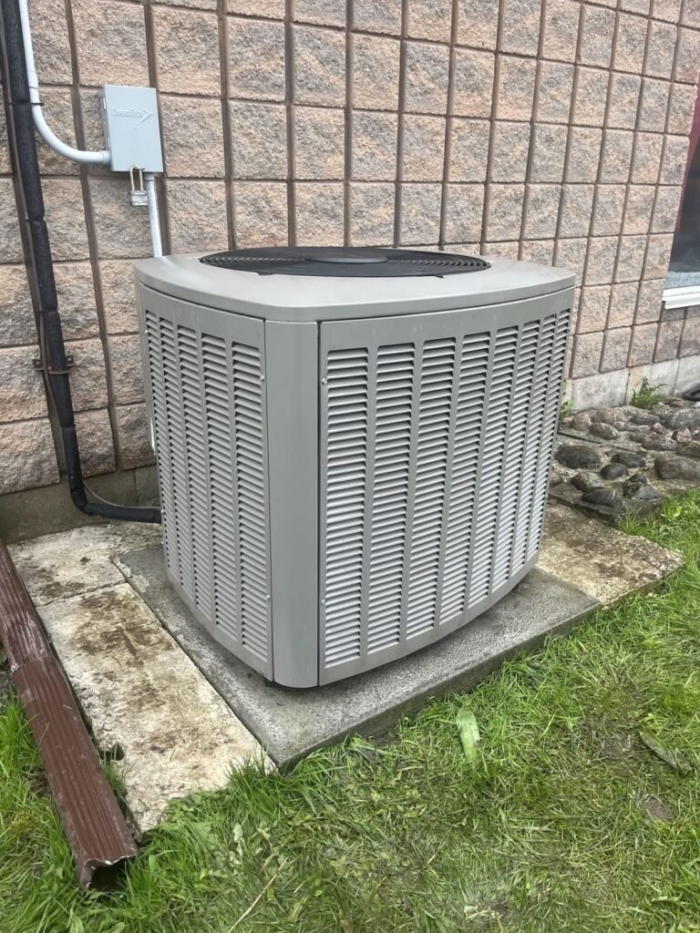 Central air conditioner outdoor condenser on a concrete pad beside a pink brick wall with an electrical disconnect box and downspout