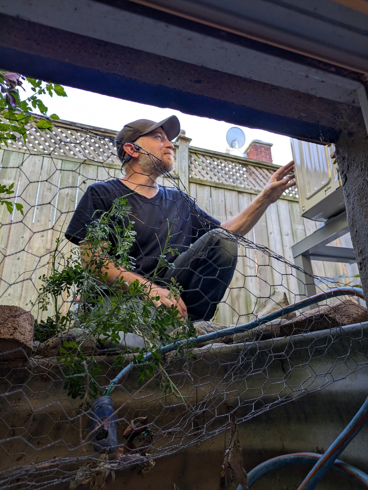 Ben Senior, Ozone Climate head technician, crouching in a crawlspace window well beside a wall-mounted mini split outdoor unit