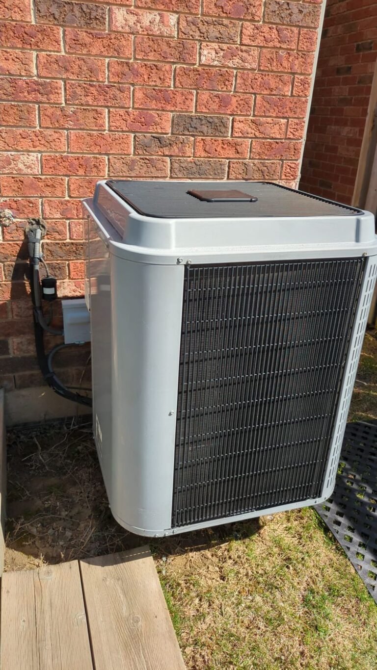 Light grey central air conditioner condenser installed beside a red brick wall with a wooden walkway and grass around it