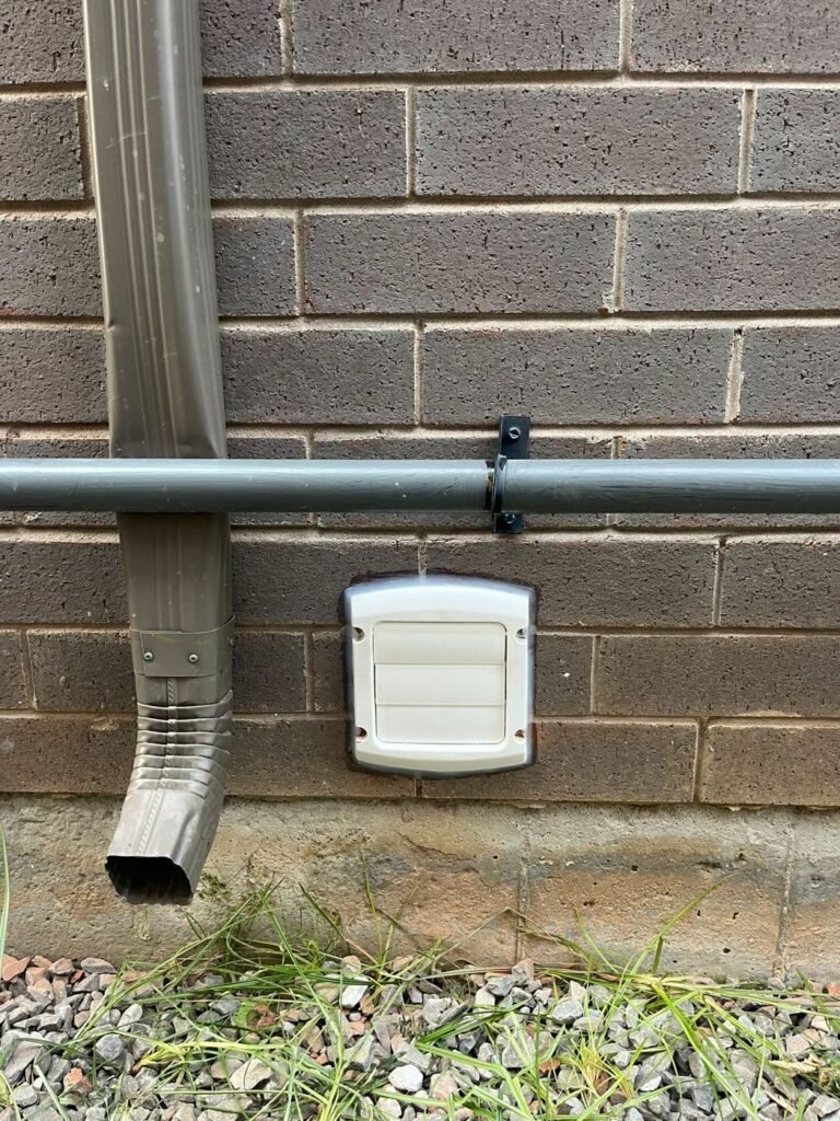 White louvered exhaust vent hood on a brick wall beside a metal downspout and grass
