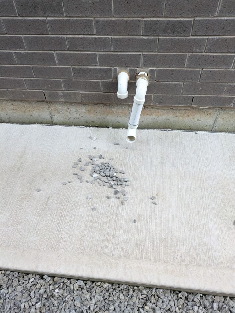 Two white PVC furnace intake and exhaust pipes passing through a dark brick wall above a concrete pad with gravel