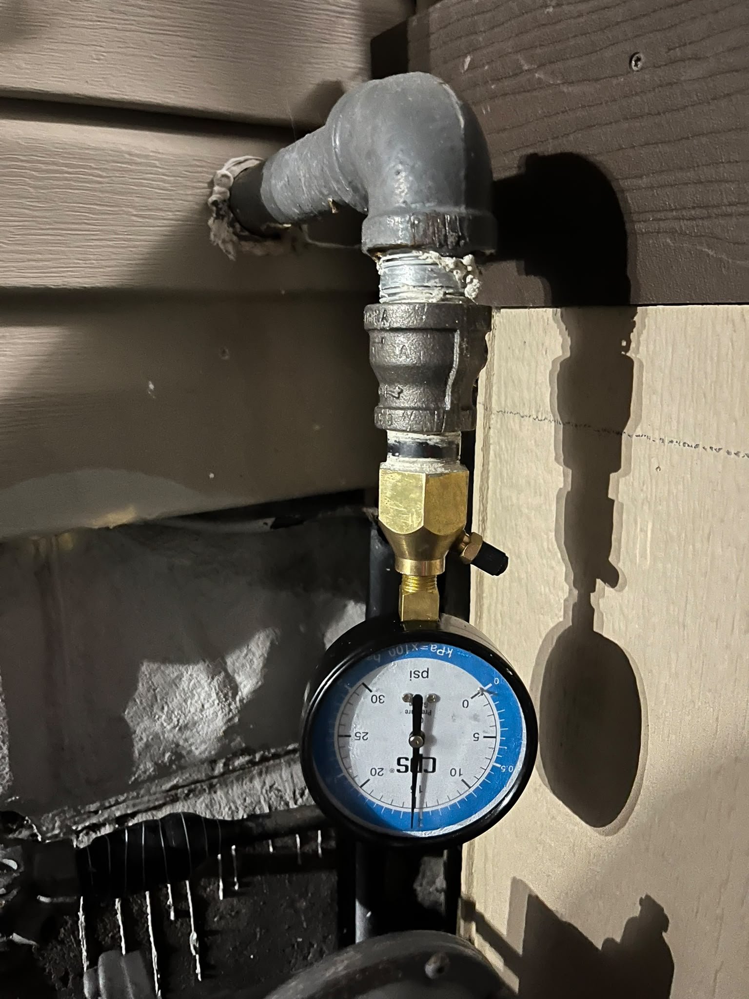 Gas line pressure test gauge in psi fitted to a black iron gas pipe elbow on an exterior wall