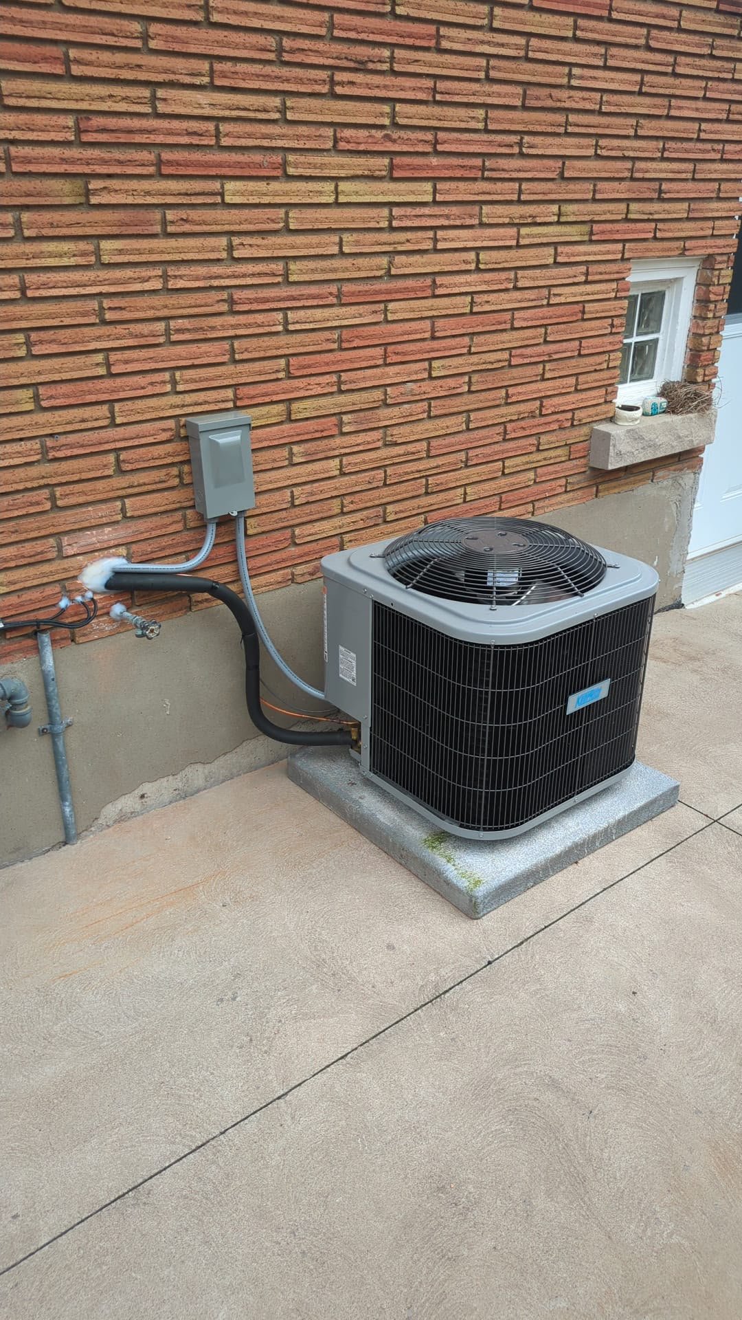 Newly installed KeepRite air conditioner outdoor unit on a concrete pad against a red brick wall with insulated line set and disconnect box