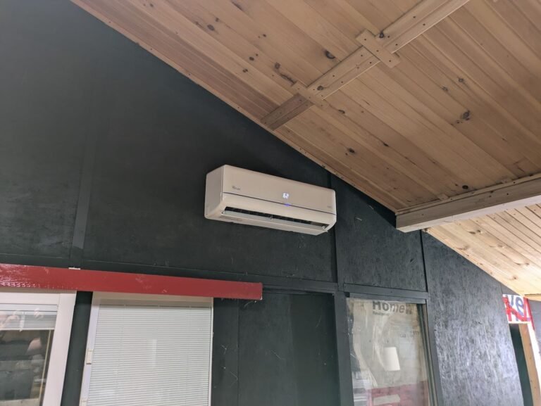 White mini split indoor unit mounted high on a dark wall below a pine plank ceiling in a garage or workshop