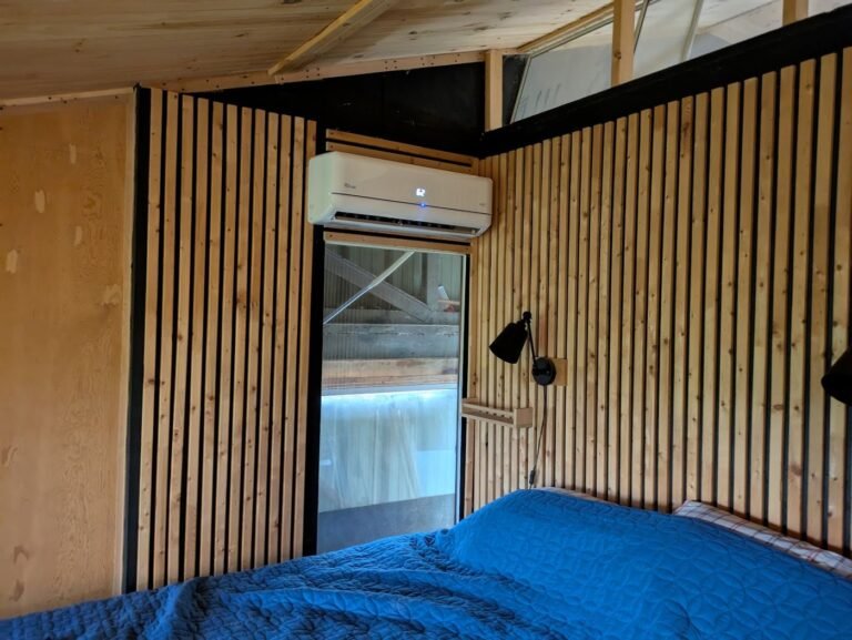 Wall-mounted mini split indoor unit above a doorway in a bedroom with vertical wood slat walls and a blue bed