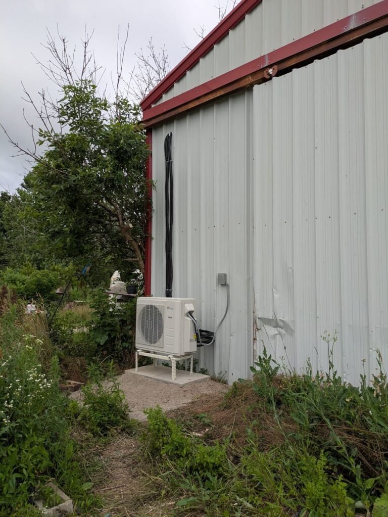 Mini split outdoor unit mounted on a concrete pad beside a metal-clad shed with a conduit running up the wall