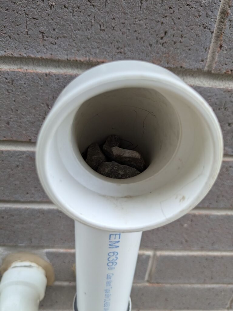 Close-up of a white PVC vent pipe terminal with gravel inside against a grey brick wall
