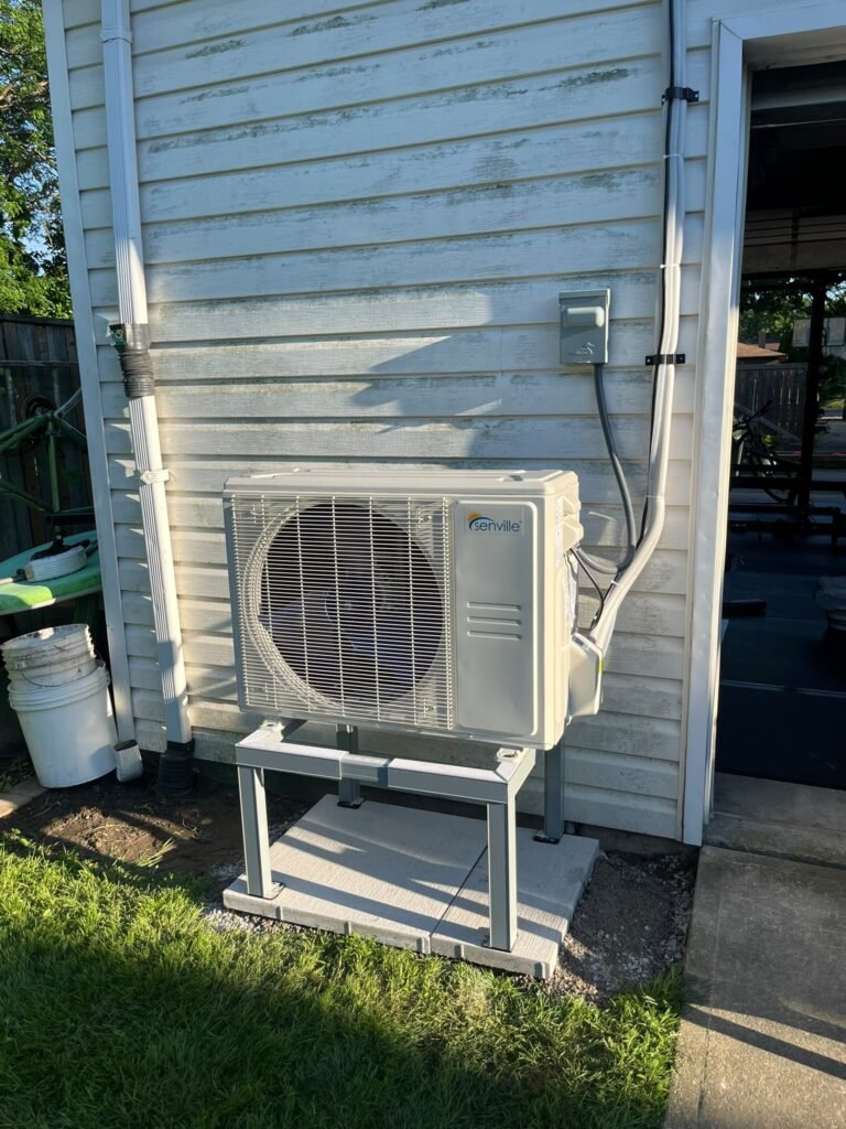 Senville mini split outdoor unit mounted on a steel stand over concrete pavers beside a white vinyl-sided wall with electrical disconnect