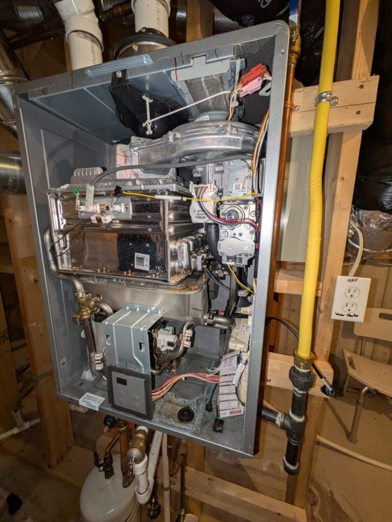 Tankless gas water heater with the front cover removed showing the burner, heat exchanger, wiring and gas pipe on a basement wall